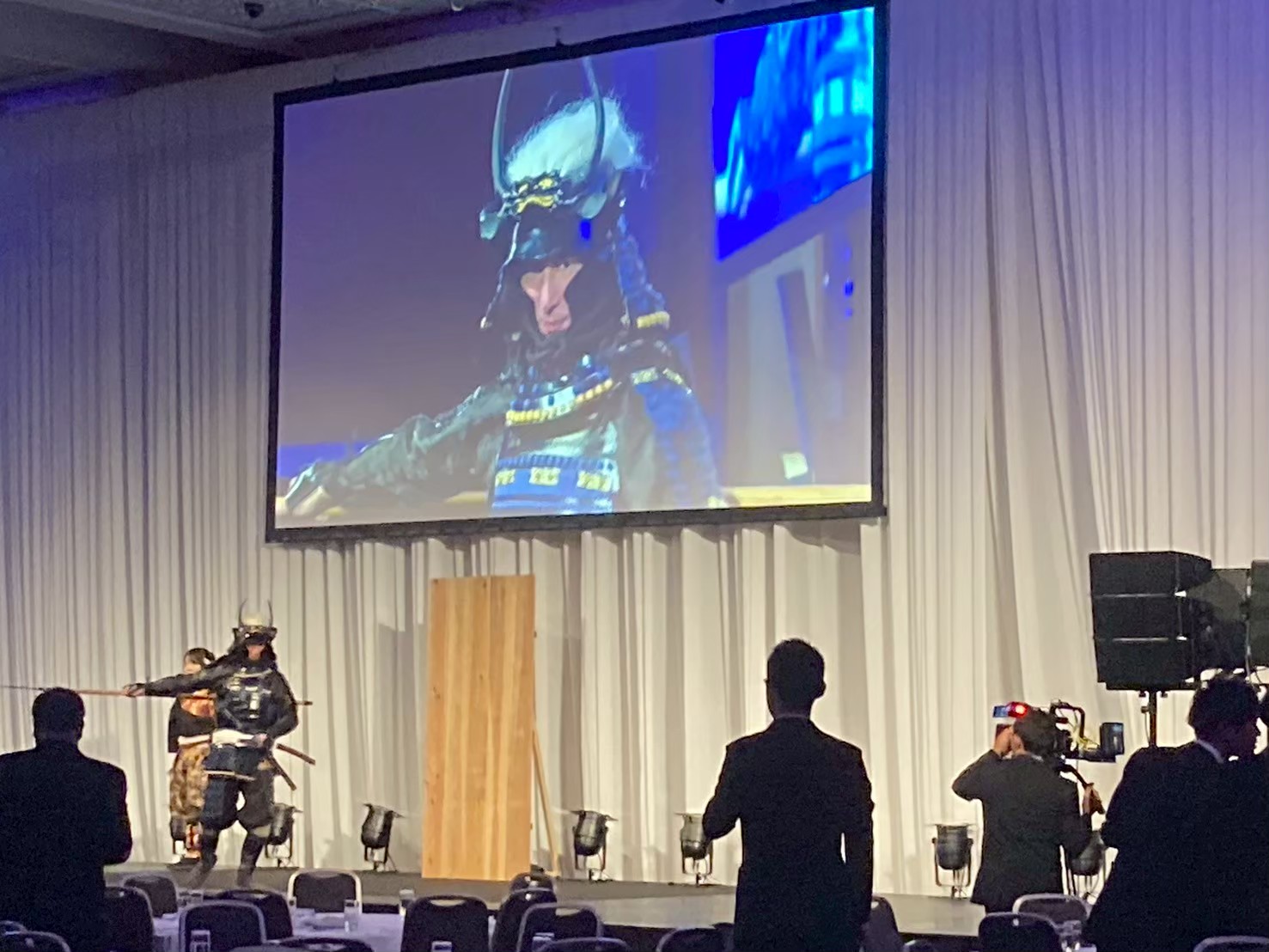 ステージ中央で甲冑姿の武将が槍を構えて登場し、その様子が大型スクリーンに映し出されている場面